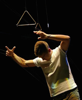 A white man in a white t-shirt grasps ahold of threads suspending musical triangles in the air