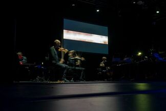 Musicians perform on a dark stage in front of an abstract video projection.
